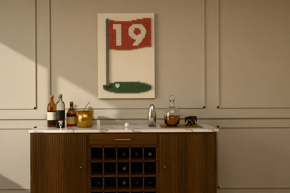 A golf-tee picture of a red pin flag numbered 19 over a green with a cup and a ball, hung tall on a light greige paneled wall above a walnut bar cabinet with a marble top, wine bottles in its cubbies, spirits, a brass ice bucket and a crystal decanter.